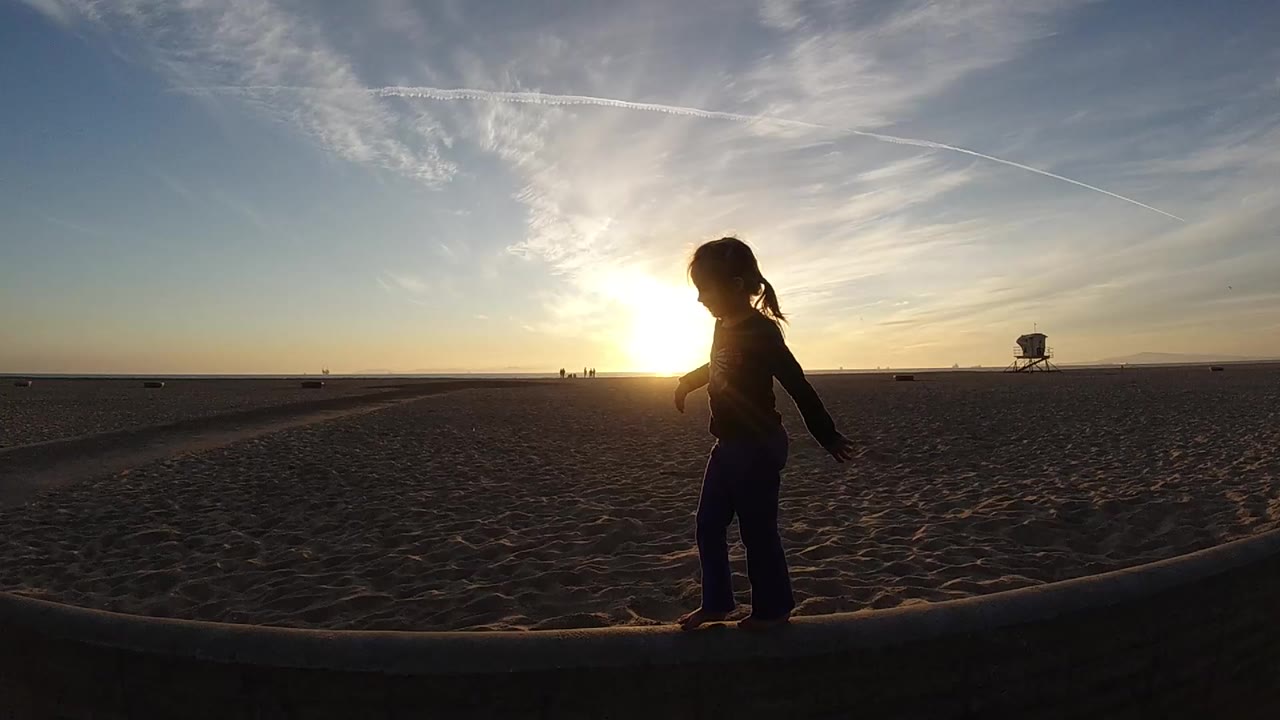 At Bolsa Chica, California, a silhouetted person walks along a low beach ledge with the sun low behind them and a lifeguard tower far on the horizon.