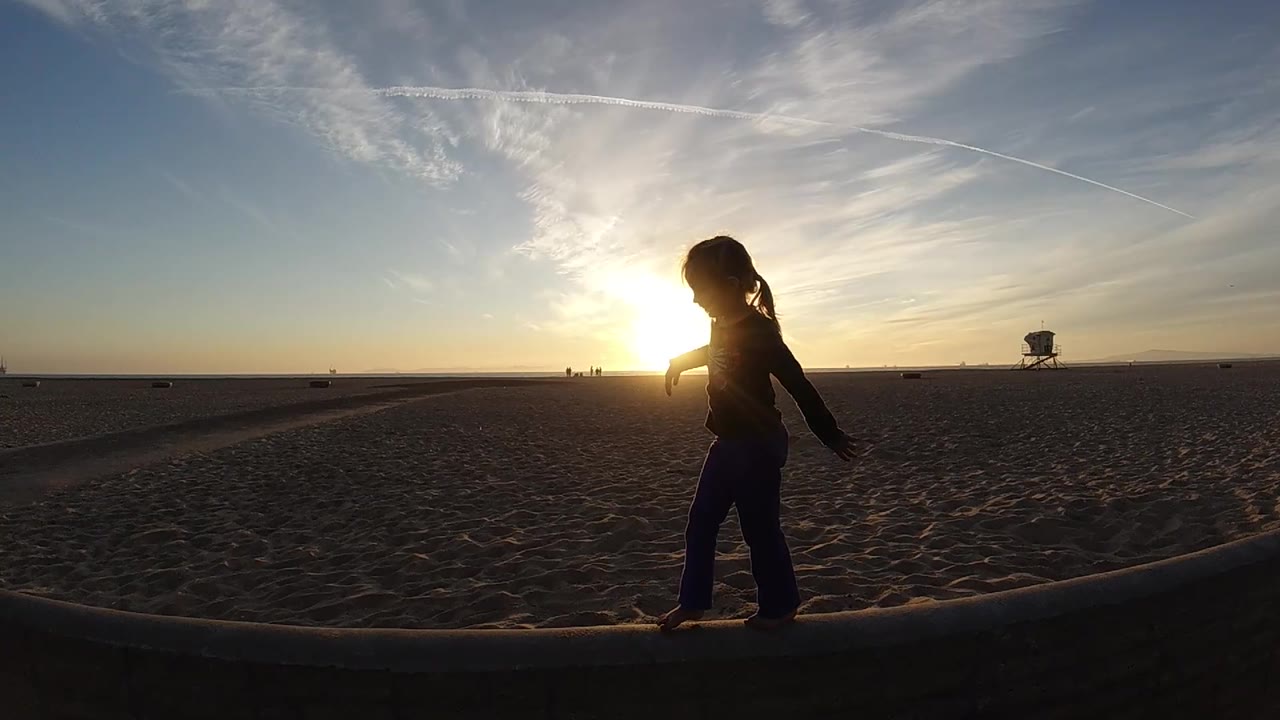 At Bolsa Chica, California, the silhouetted person continues along the ledge near the right side of the frame as the distant lifeguard tower remains visible over the sand.