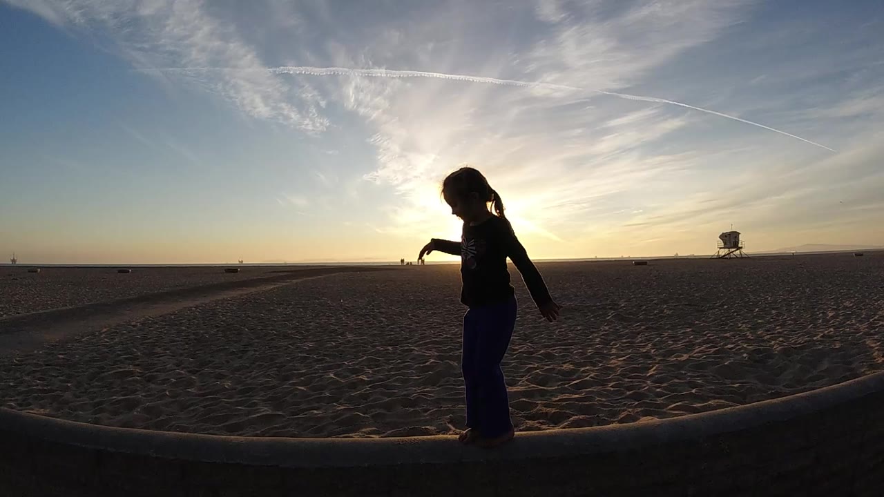 A silhouetted child stands on a low curb at Bolsa Chica, California, with the sun behind them, rippled sand ahead, and a lifeguard tower near the horizon.