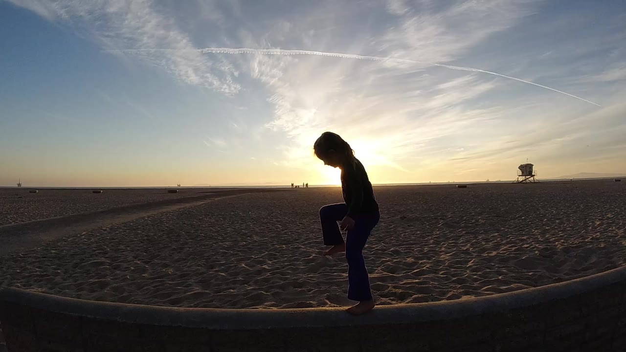 In Bolsa Chica, California, a silhouetted child lifts one knee on a low concrete edge with open sand, sunset glare, streaked clouds, and a distant lifeguard tower behind her.