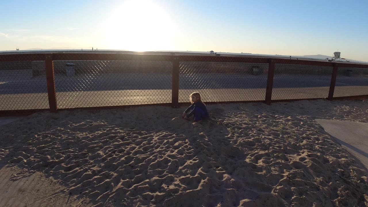 Child playing in sand beside a fence with the sun low over the horizon.