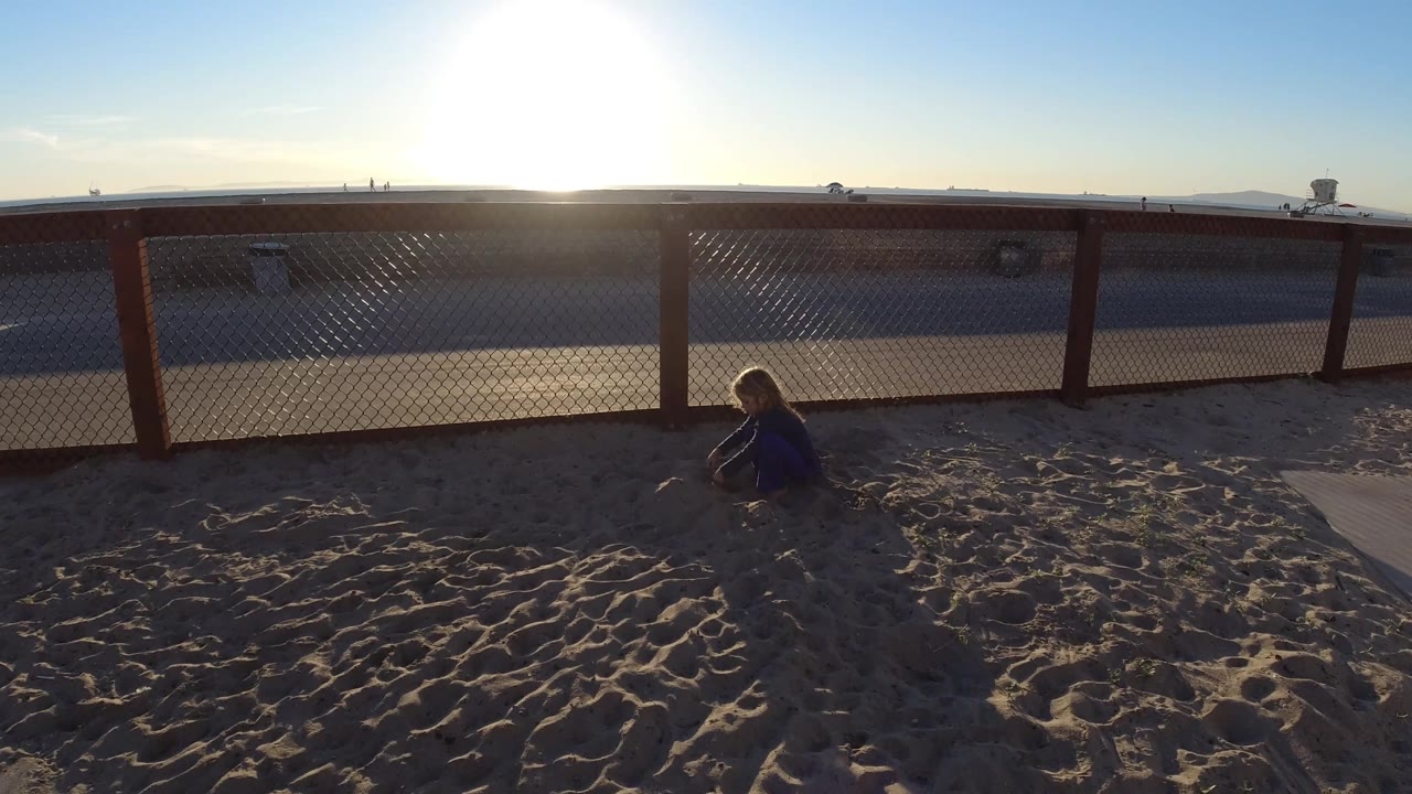 At Bolsa Chica, California, the child appears nearer the right side of the sandy foreground, with the fence line and blown-out sun centered above the beach.