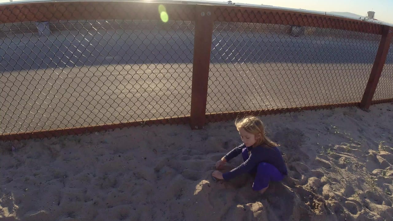 At 0:00 in Bolsa Chica, California, a young blonde girl crouches low in pale sand near a chain-link fence under strong side light.