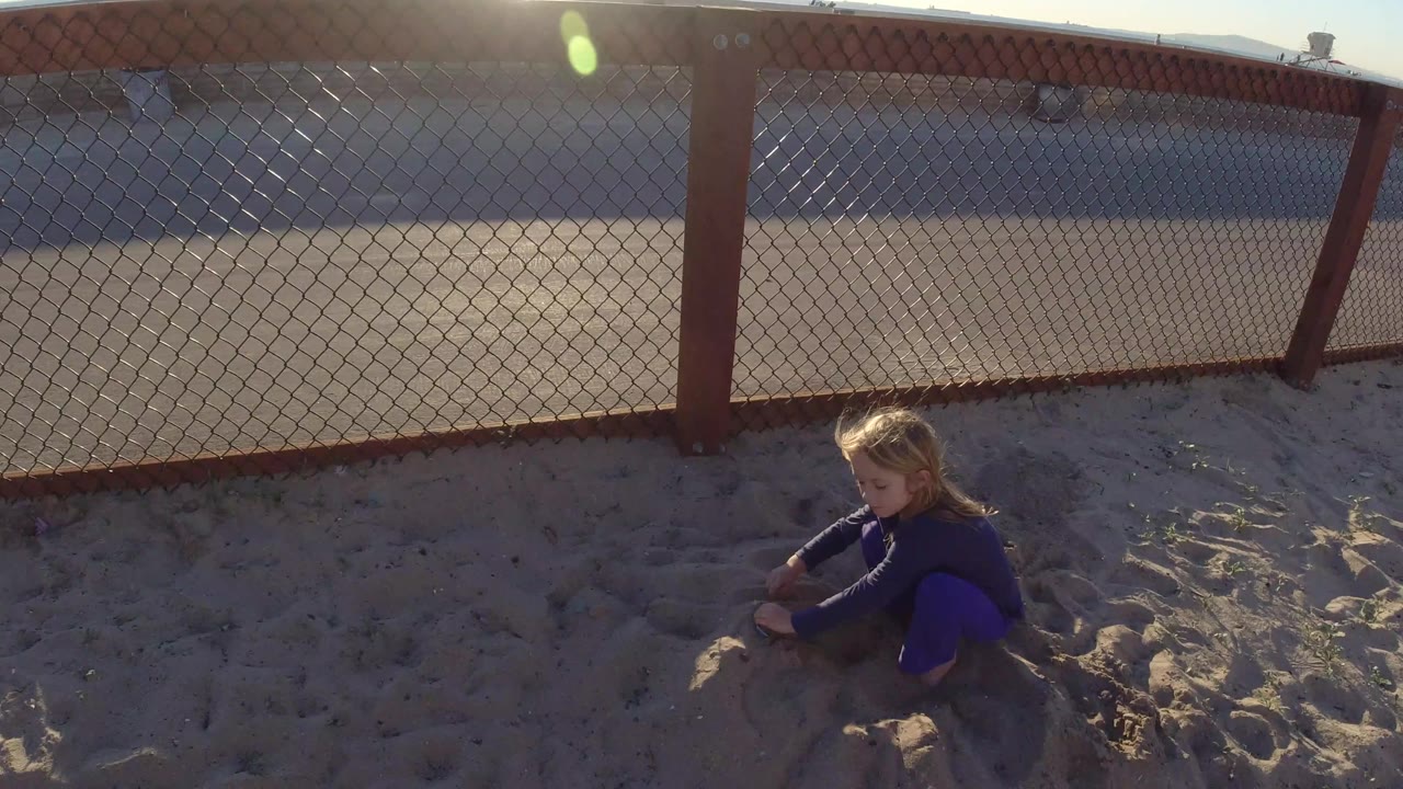Young girl kneeling in sand with fence behind her.