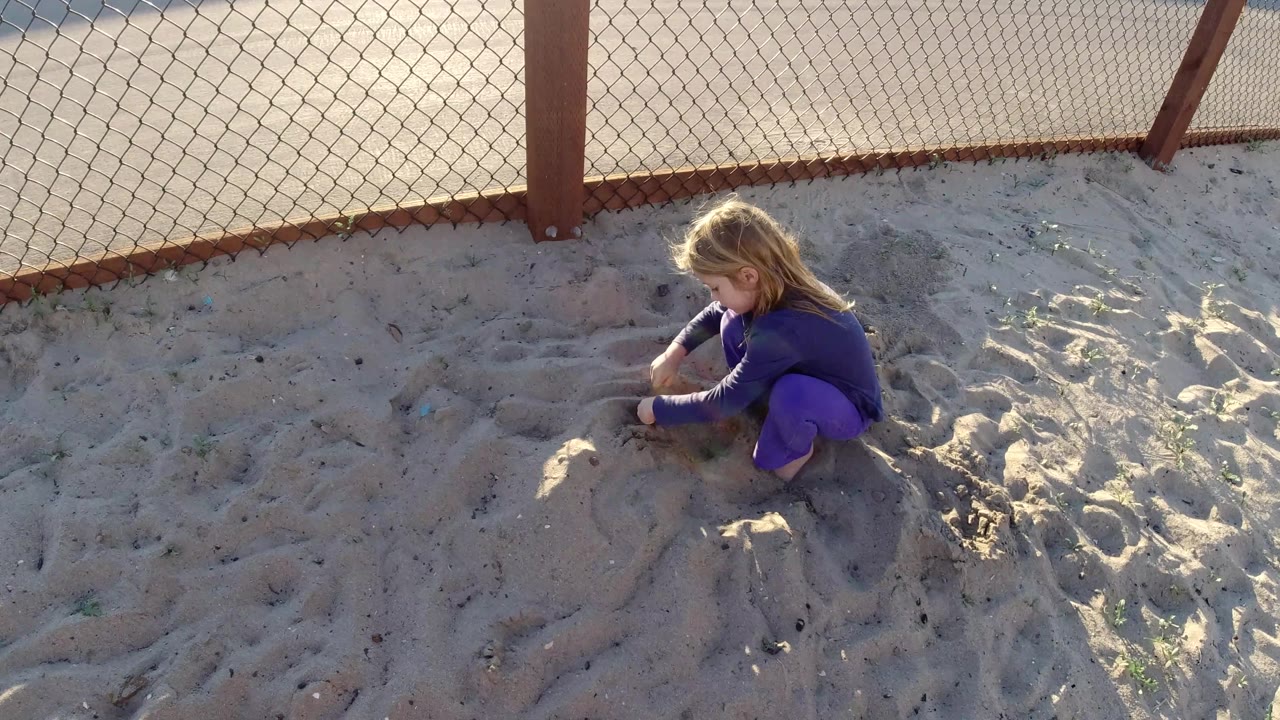 At 0:02 in Bolsa Chica, California, the girl sits farther to the right of frame, looking down at the sand with bright sun catching her hair.