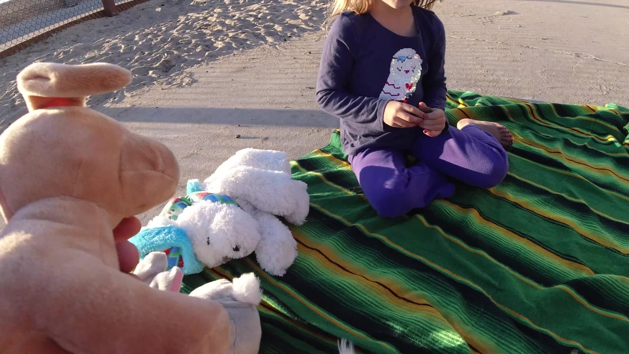 Hand holds a tan plush toy toward the child on the blanket.