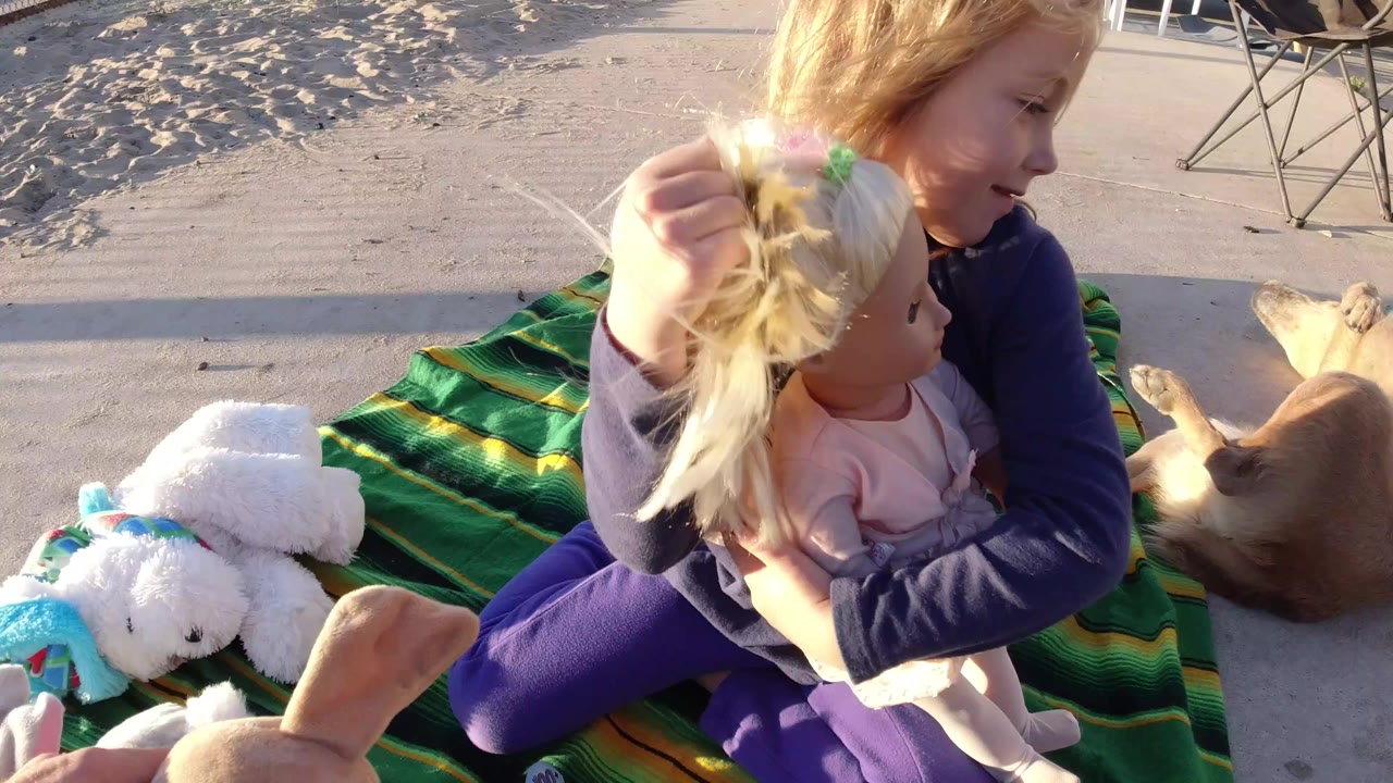 At Bolsa Chica, California, a young blonde girl sits on a green striped blanket holding a blonde doll while a small tan dog lies close to her side.