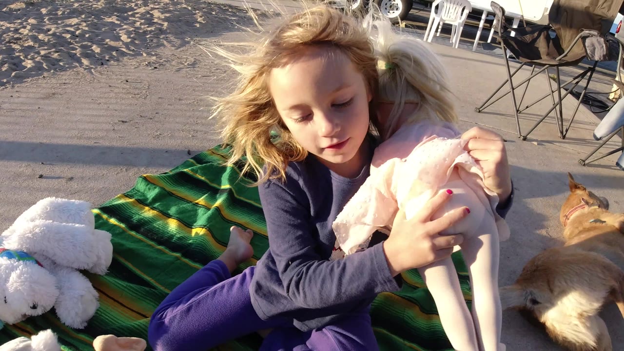 In Bolsa Chica, California, the girl turns more toward the camera while holding the pink-clothed doll, with wind in her hair and the tan dog partly visible beside the blanket.