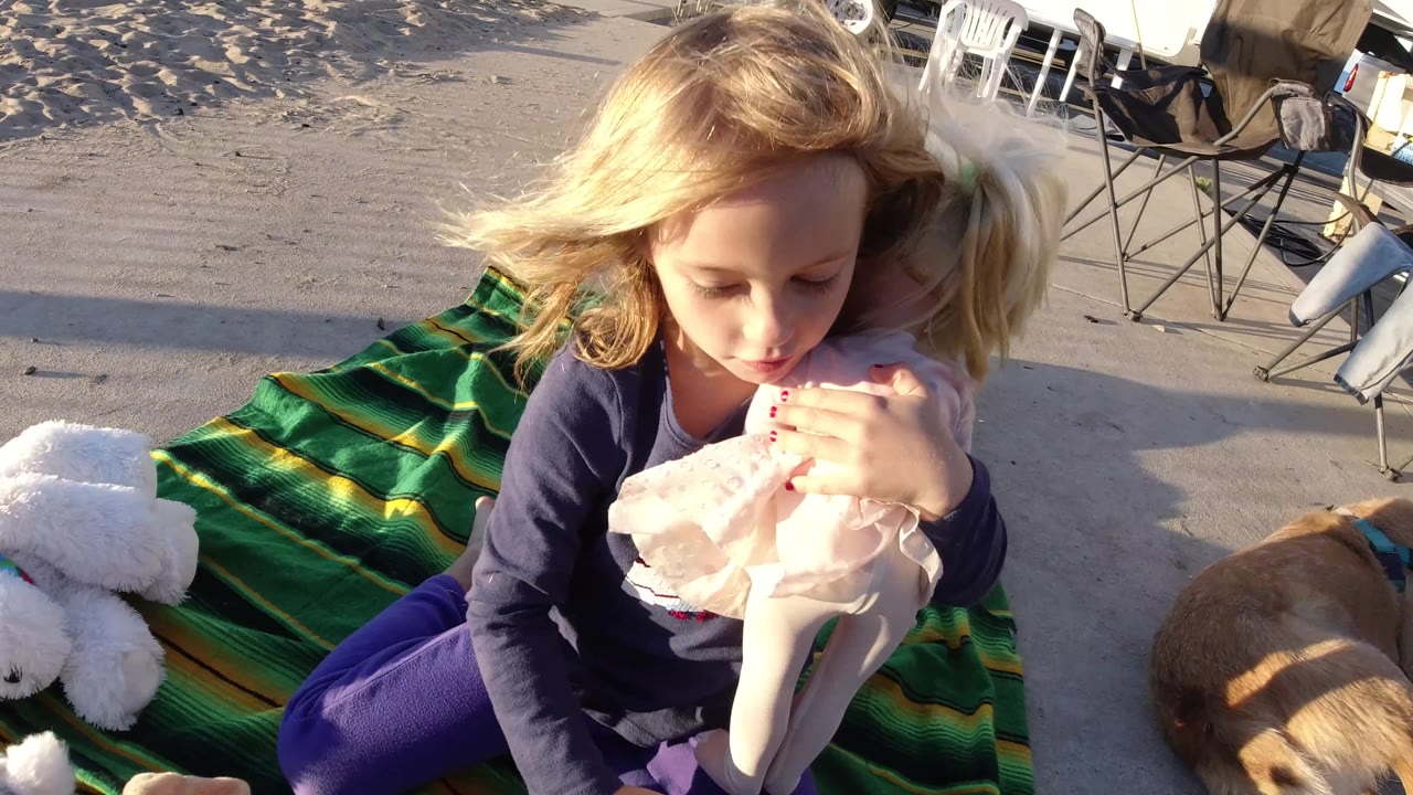 Girl kisses a doll while seated on a green blanket outdoors.