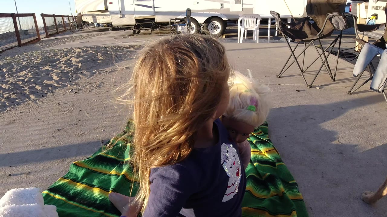 A young girl in Bolsa Chica, California sits on a green striped blanket holding a small light-colored dog, with trailers and folding chairs behind her.