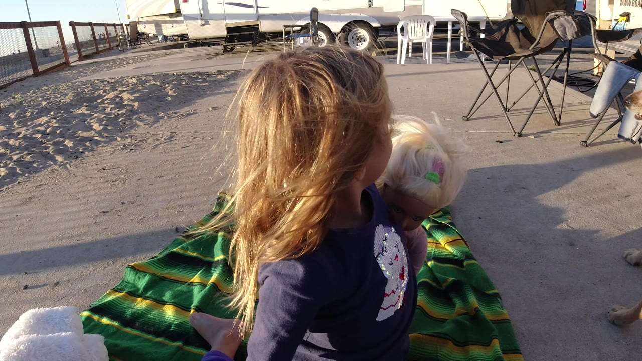 The child and small dog stay close together on the blanket in Bolsa Chica, with long shadows, RVs, chairs, and sandy ground visible around them.