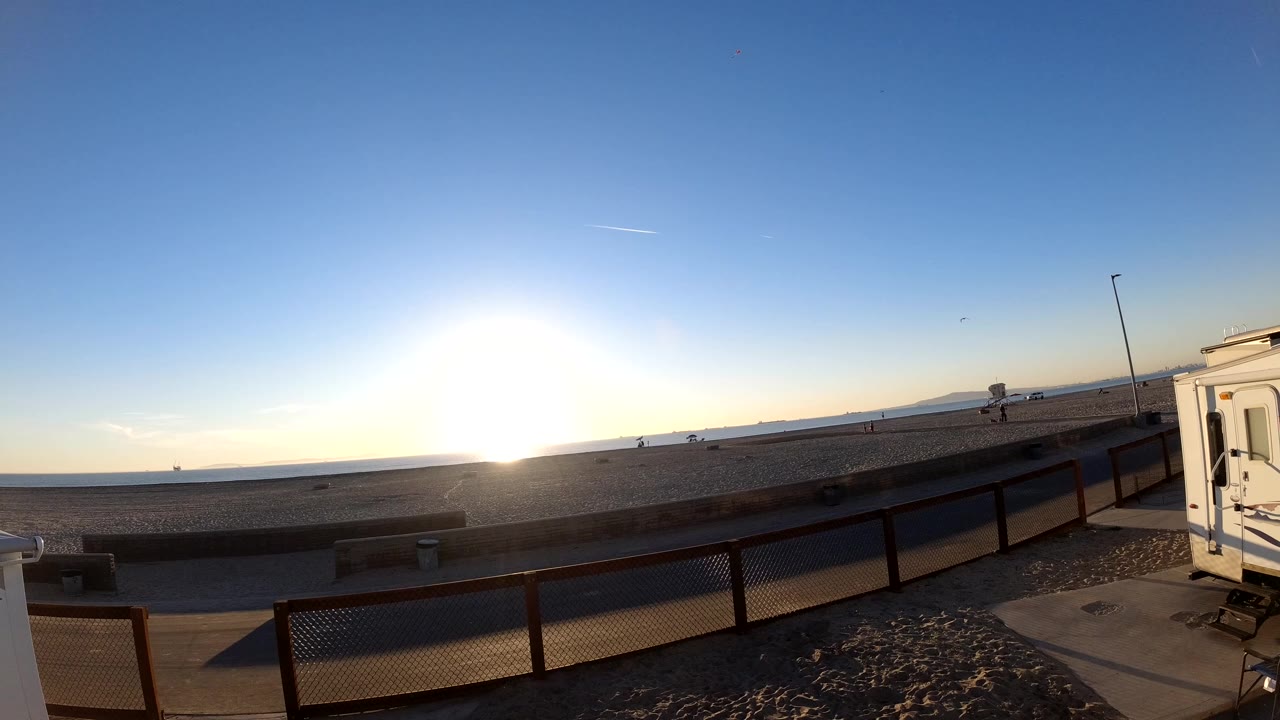 Bolsa Chica coastal road and beach foreground under a bright low sun, with railing, ocean horizon, and no clearly visible people.