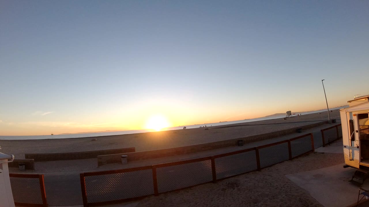 At Bolsa Chica, California, the sun sits low on the ocean horizon beyond a wide beach, with a red foreground fence, paved path, small white structure at right, and no visible people.