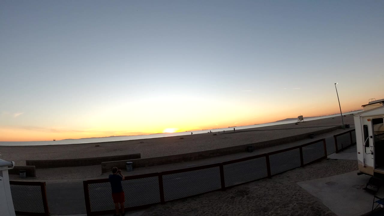 Bolsa Chica, California sunset at 0:00 with the sun low on the horizon, a dark foreground railing, a small structure at right, and a silhouetted person near the lower left.