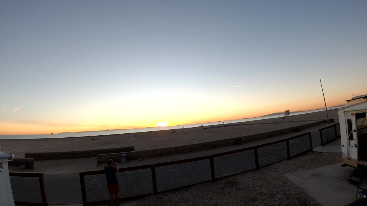 Bolsa Chica, California sunset at 0:01 with the bright horizon centered, dark fence lines across the foreground, a silhouetted person near the lower left, and a small building edge at right.