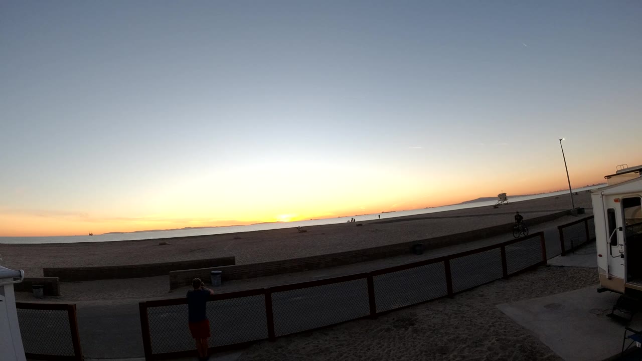 Bolsa Chica, California sunset at 0:02 with a bright orange horizon, dark fence in the foreground, a small structure at the right edge, a foreground silhouette, and a distant person on the path.