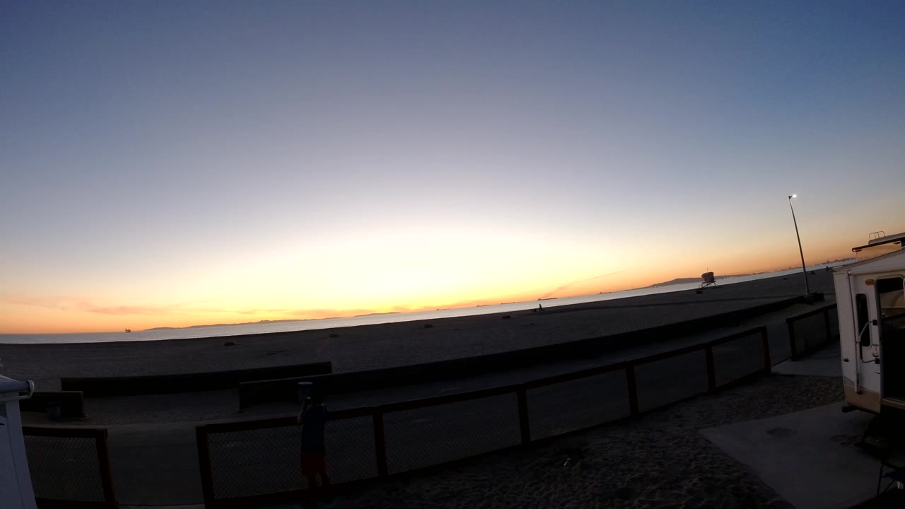 Early frame of a Bolsa Chica sunset with an orange horizon, dark concrete foreground, a railing, a right-edge structure, and no visible people.