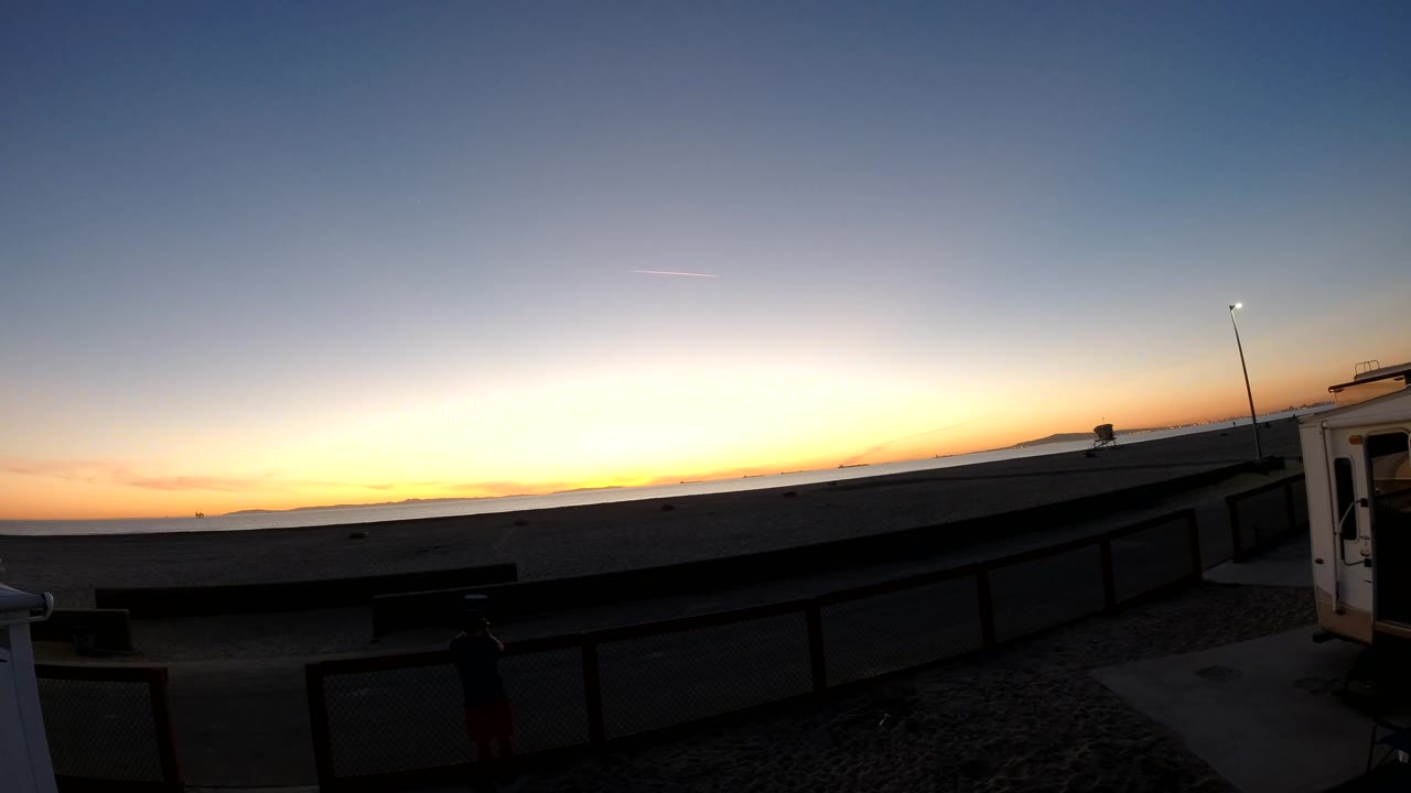 Later Bolsa Chica sunset view with a brighter orange horizon, silhouetted railing, a lit vertical pole near the right side, and no visible people.