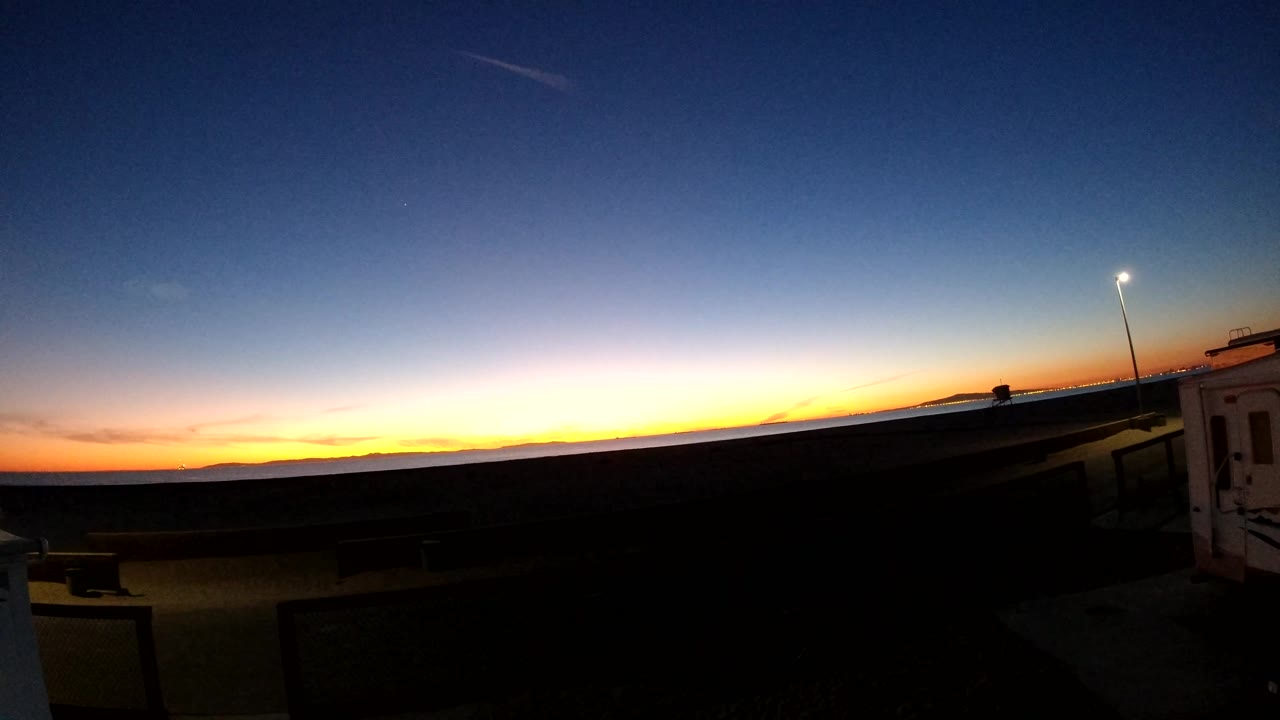 Bolsa Chica, California sunset frame with the foreground nearly black, a narrow orange horizon, a lit street light near the right edge, and no visible people.