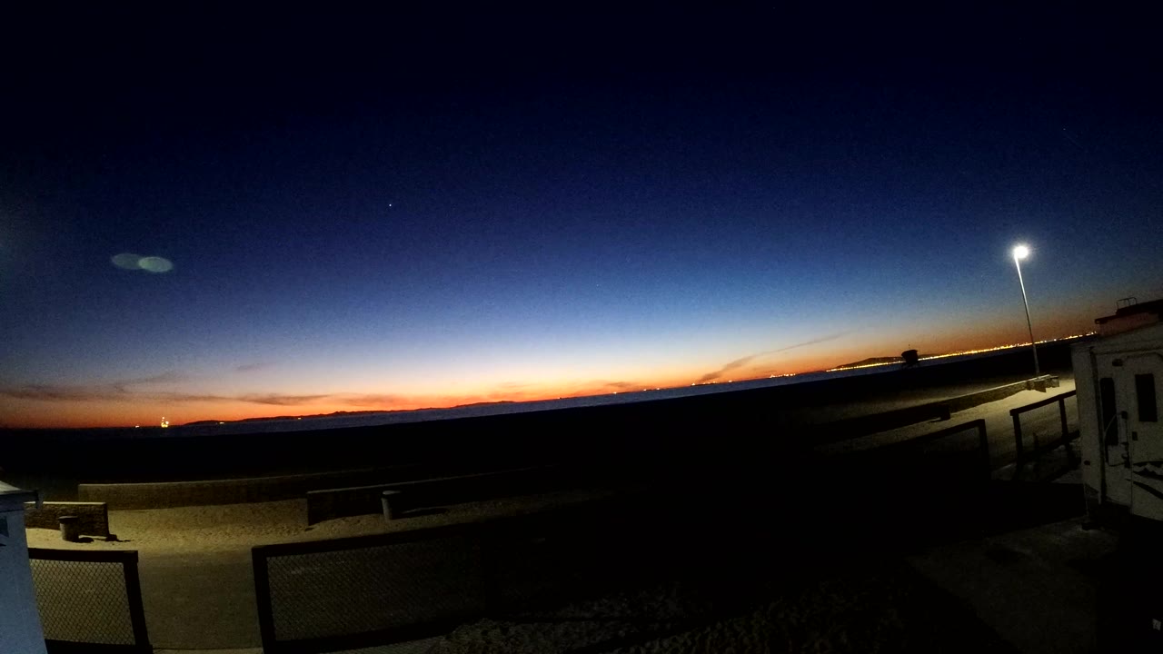 A later Bolsa Chica sunset view shows the blue sky deepening above the orange horizon, with the park foreground mostly dark, a right-side lamp pole, and no visible people.