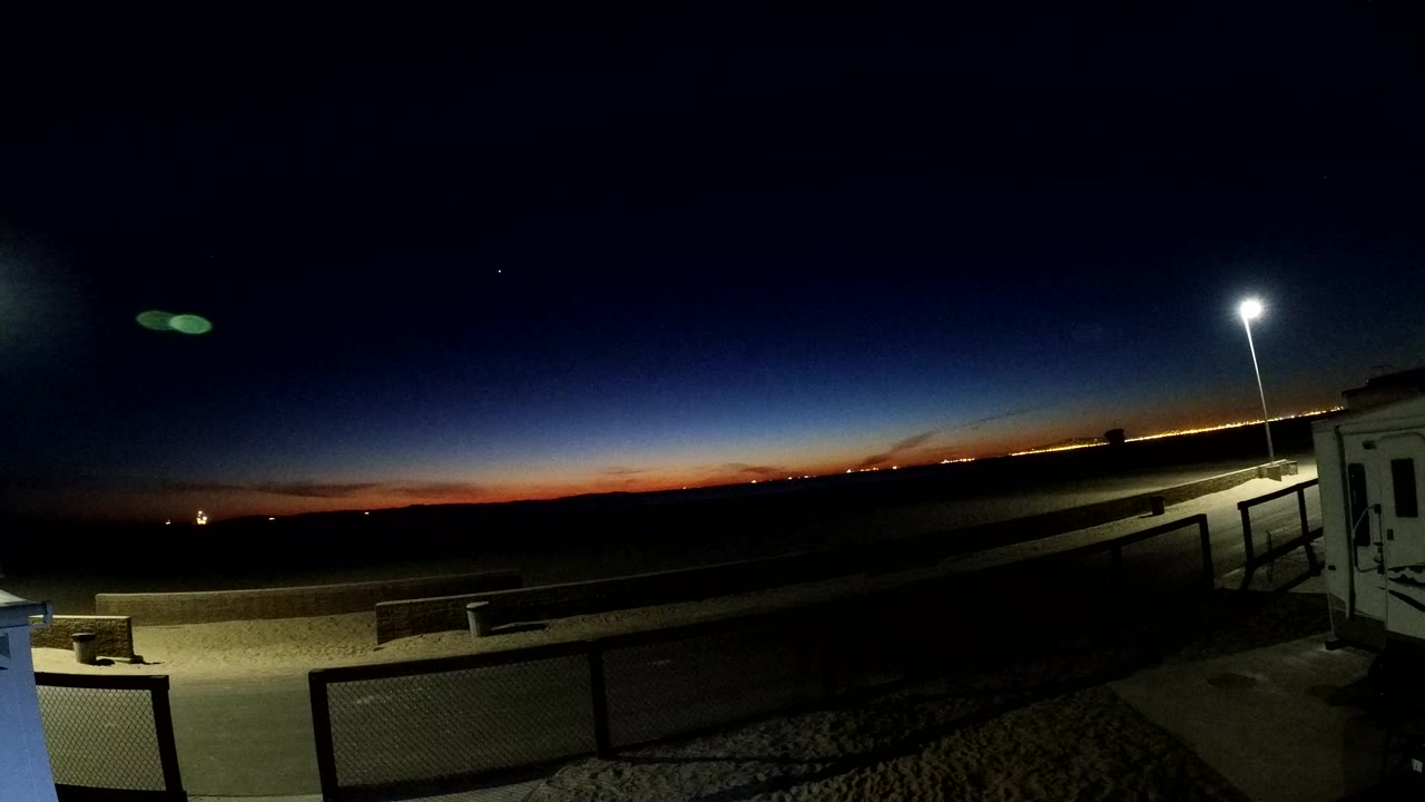 At Bolsa Chica, California, the opening frame shows a dark sky with a thin orange sunset band, railings in the foreground, a bright pole lamp at right, and no clearly visible people.