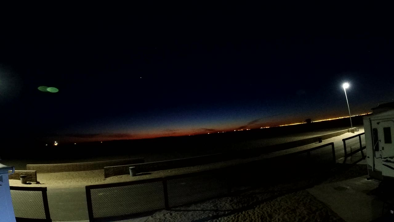 Twilight sky over railings, pavement, and a bright pole lamp at the coast.