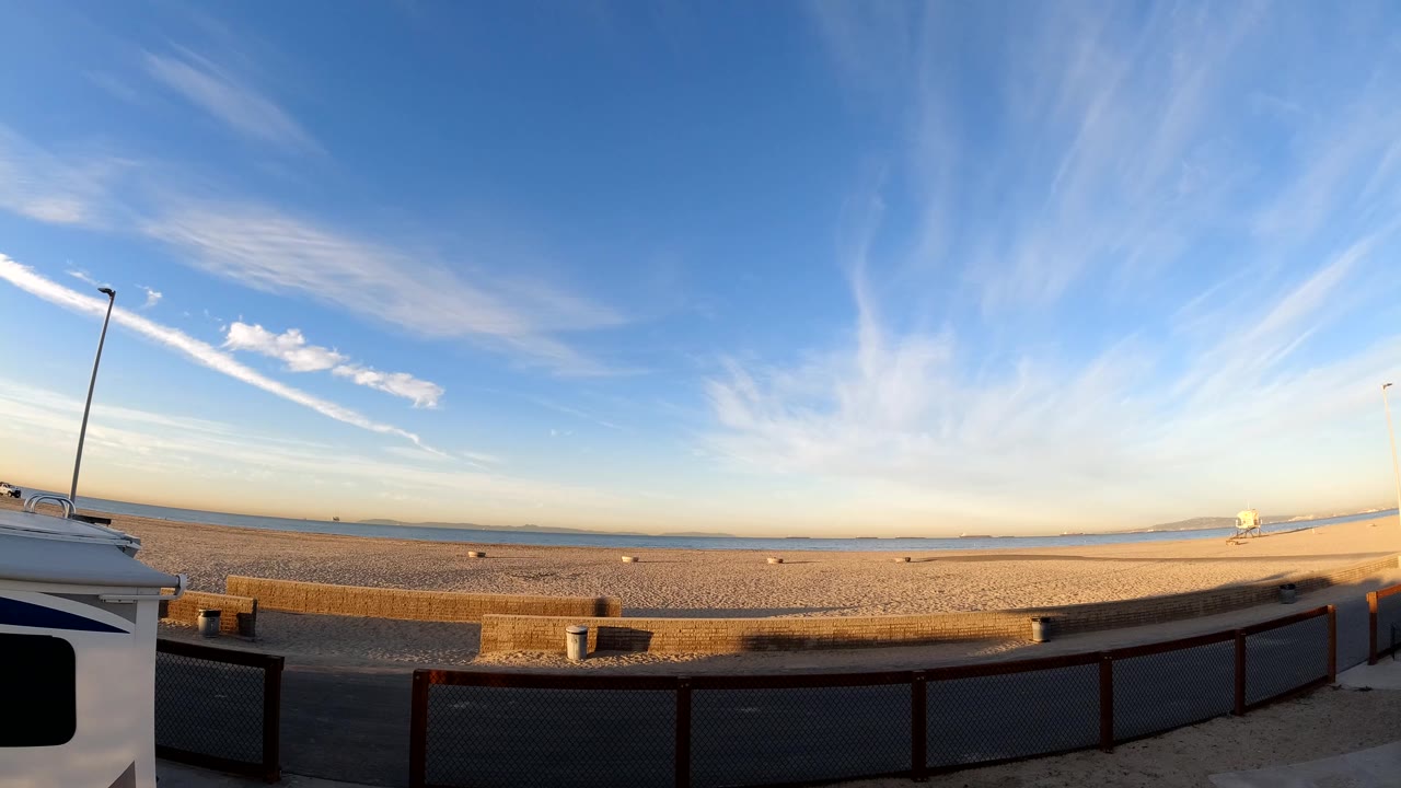 Bolsa Chica, California frame with wispy clouds stretching across a blue sky above sand, a low water horizon, foreground railing, and no visible people.