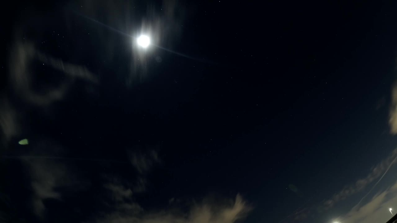 Bright sun in a dark sky with thin clouds and lens flare.