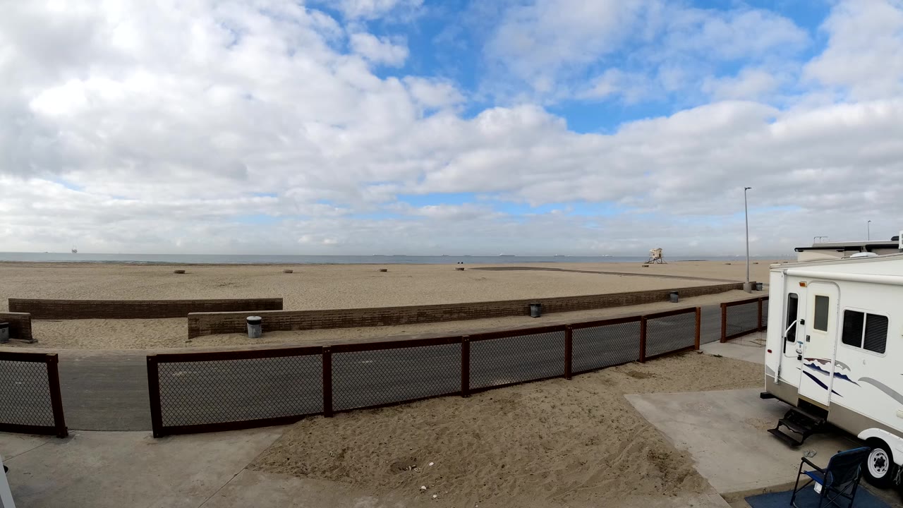 Wide Bolsa Chica beach view with sandy foreground, fenced promenade, ocean horizon, layered clouds, and a white RV at the right edge with no visible people.