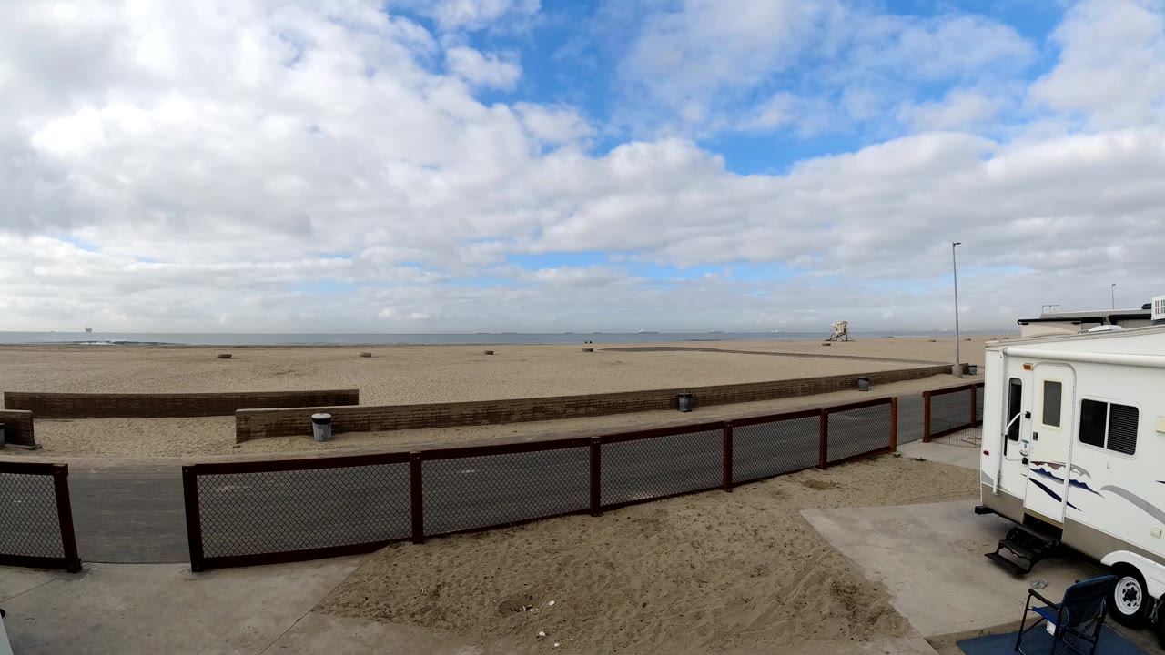 Bolsa Chica beach scene with the RV more centered at frame right, the fenced promenade across the midground, broad sand, distant water, and no visible people.