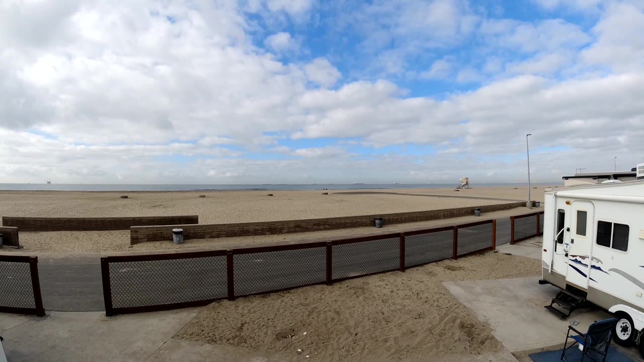 Bolsa Chica beach panorama showing the fenced walkway and low wall beneath a cloud-filled sky, with the white RV partly cropped at the right and no visible people.