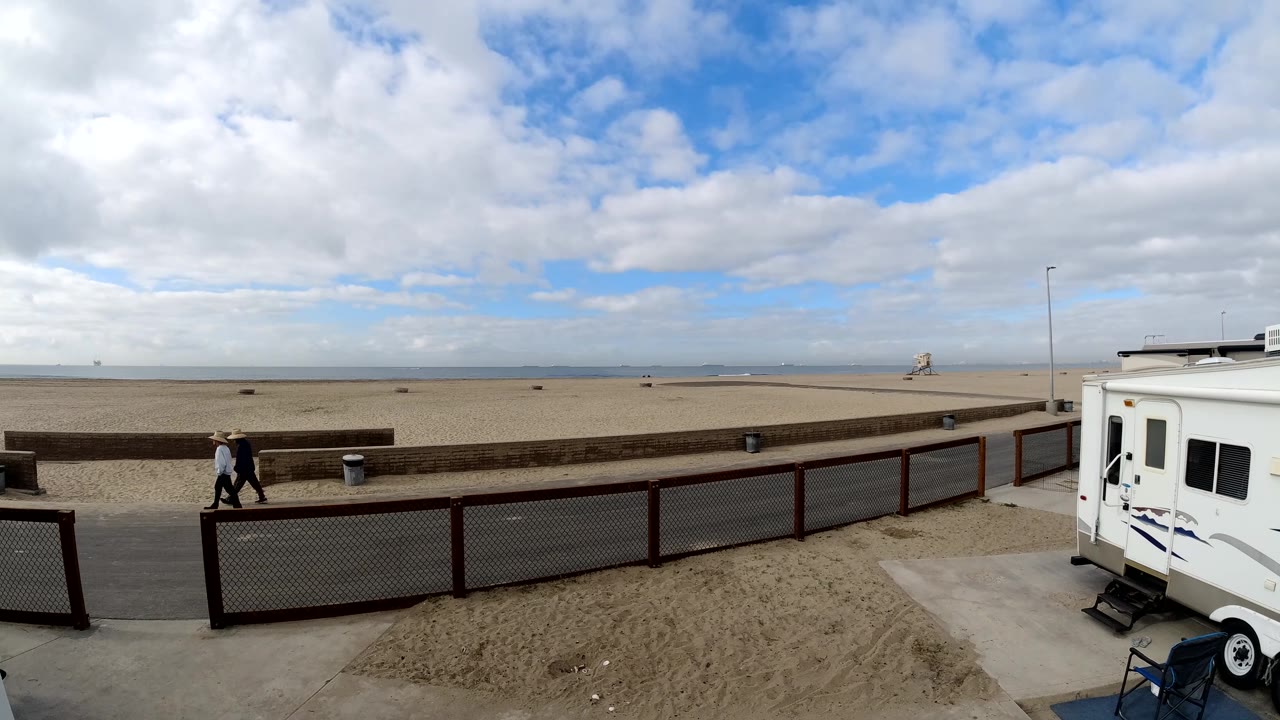 Two distant pedestrians on the promenade beside a sandy beach and parked RV.
