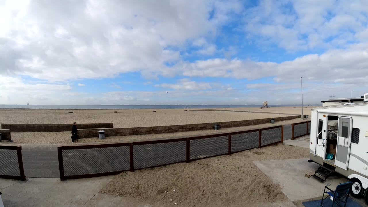 Wide elevated view in Bolsa Chica, California showing a white trailer at right, fenced path, sandy beach, distant water, cloudy sky, and one person walking near the left side.