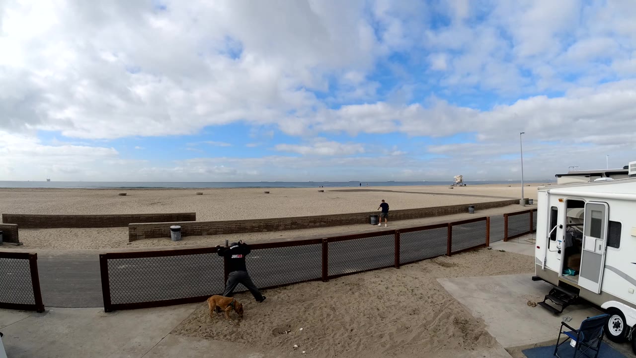 Bolsa Chica beach frame with the trailer at right, a person gesturing near the fence, a dog on the sand, another distant pedestrian, and open beach under broken clouds.