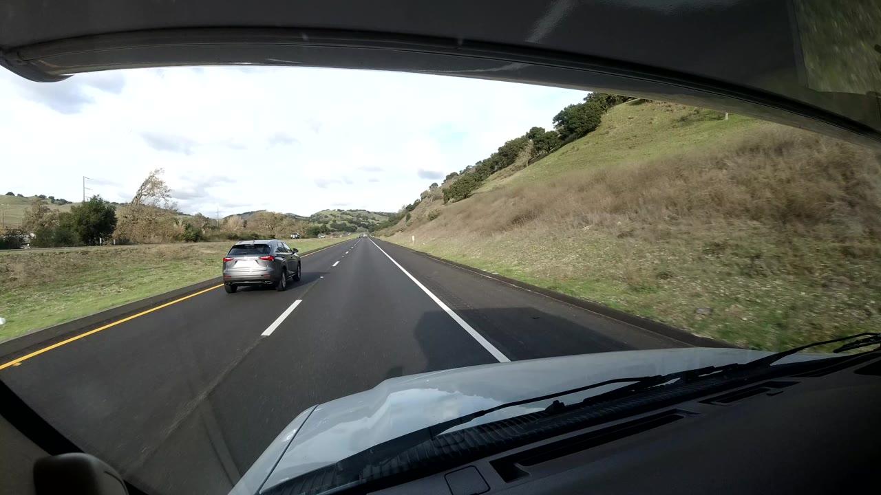Inside-vehicle view on the Coast California road with the hood at the bottom, a car ahead in the left lane, grassy hills to the right, cloudy daylight, and no visible people.