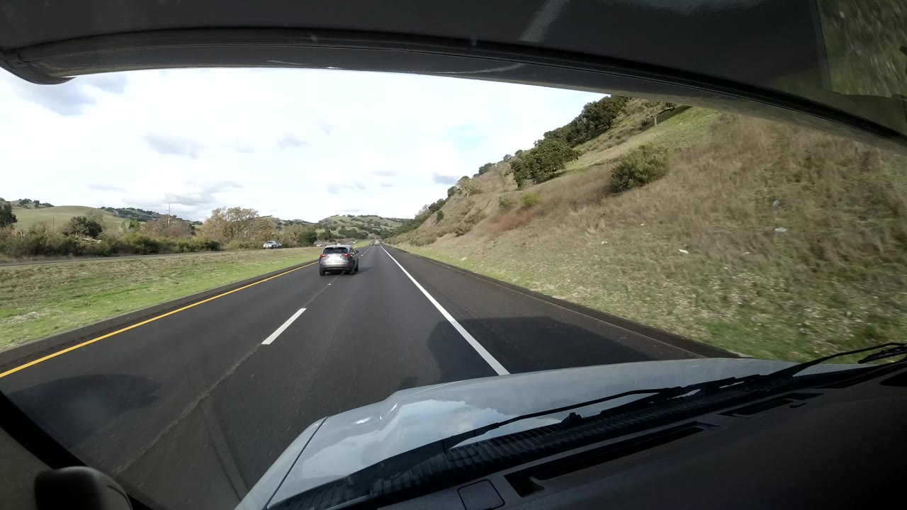 Forward dash view from inside a vehicle on the Coast California road as the car ahead appears smaller, with white lane markings, grassy shoulder, hillside, cloudy sky, and no visible people.