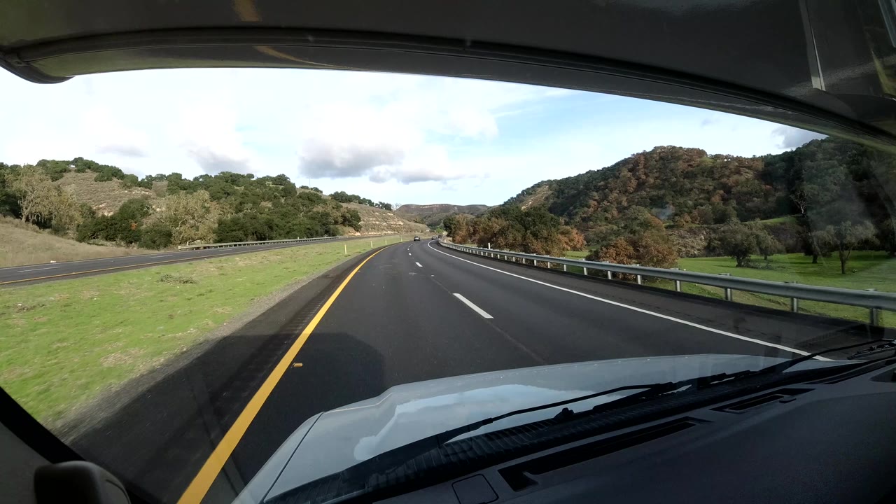Inside-vehicle view of a curving Coast California highway with a yellow left line, guardrail at right, green roadside grass, tree-covered hills, cloudy daylight, and no visible people.