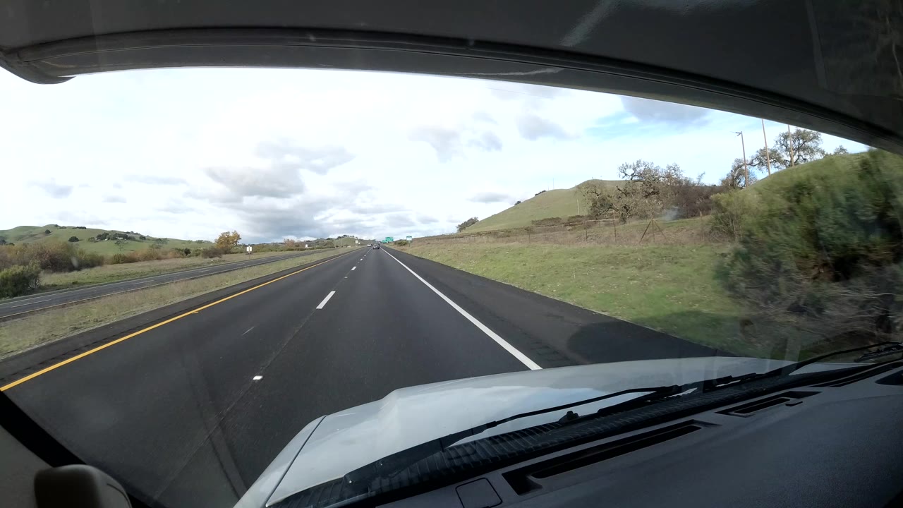 Straight multi-lane road seen through a windshield with grassy hills and open sky.