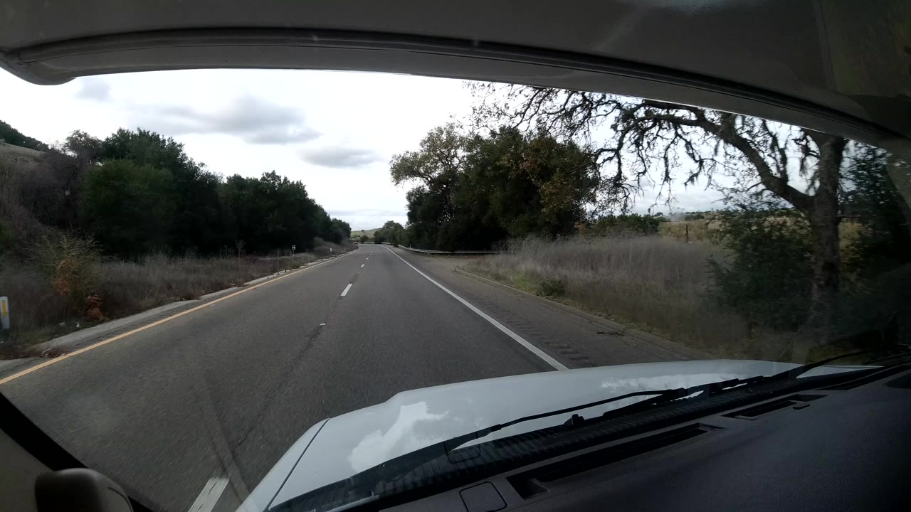 Start of a California Coast road drive seen through a windshield, with a two-lane highway, roadside trees, cloudy sky, the vehicle hood, and no visible people.
