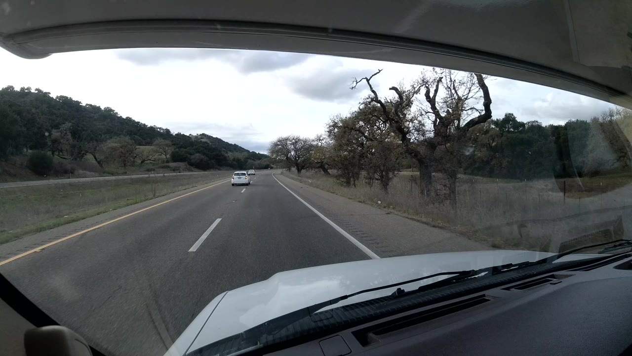 Forward windshield view on a California Coast highway with a white car ahead, bare trees to the right, low green hills in the background, cloudy daylight, and no visible people.