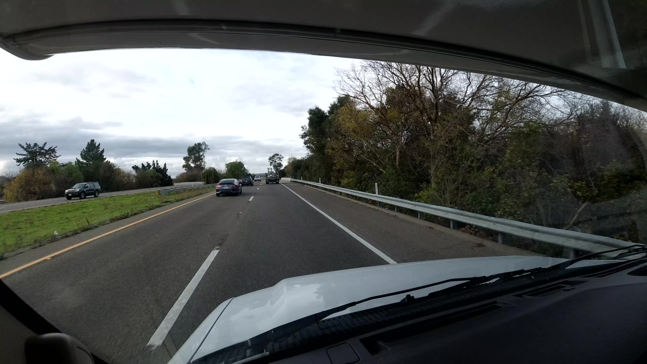 Forward vehicle view on a Coast California highway with cars ahead, guardrails, trees, overcast sky, hood and wipers in the foreground, and no visible people.
