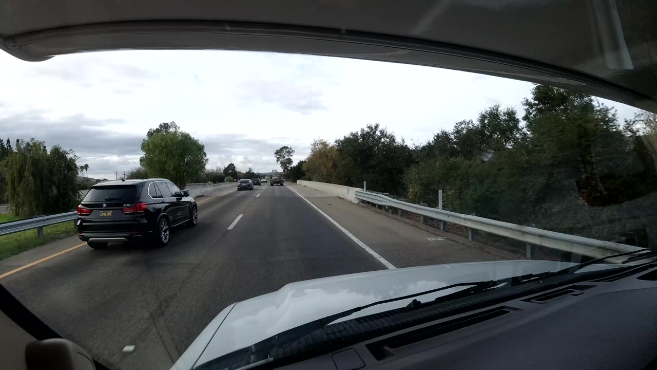A black SUV passes near the windshield-framed roadway on a Coast California highway, with other cars ahead, guardrails, trees, cloudy sky, and no visible people.