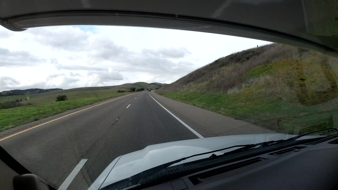 Forward dashboard view on a Central Coast California highway, with the vehicle hood at bottom, green hills ahead, cloudy daylight, and no visible people.