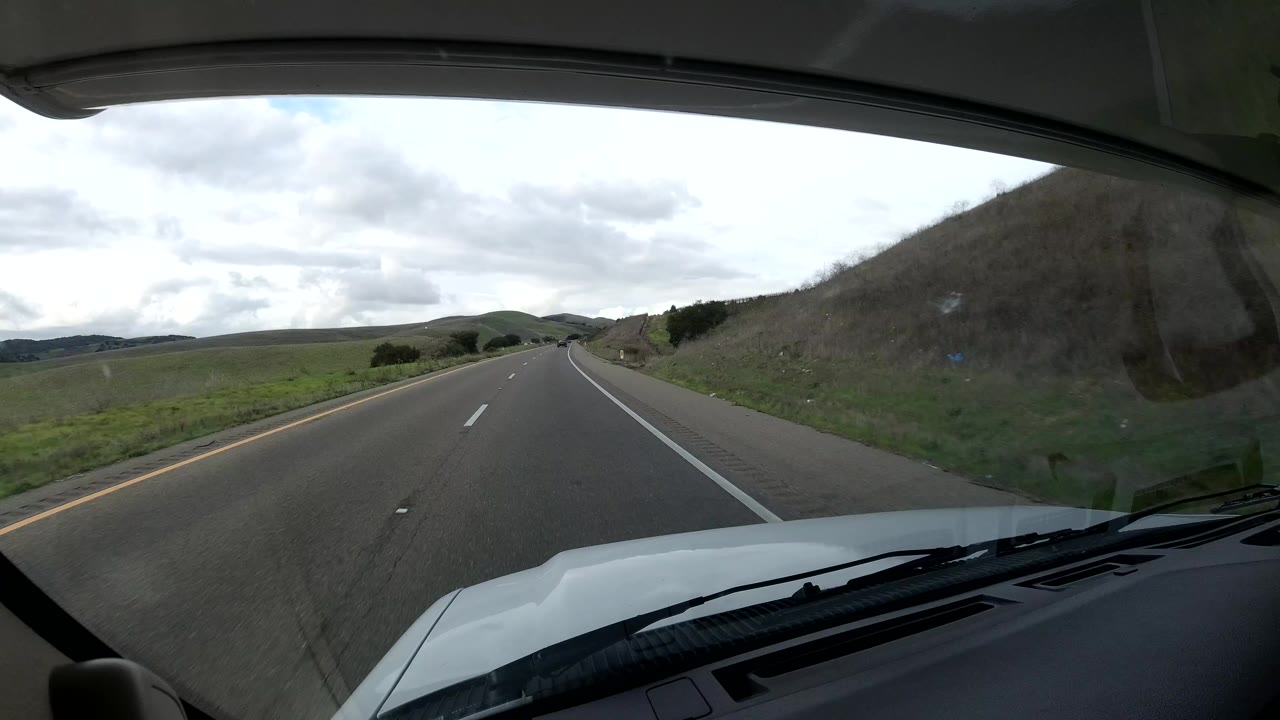 Central Coast California road view from inside the vehicle, with a hillside rising beside the right shoulder, open lanes ahead, and no visible people.