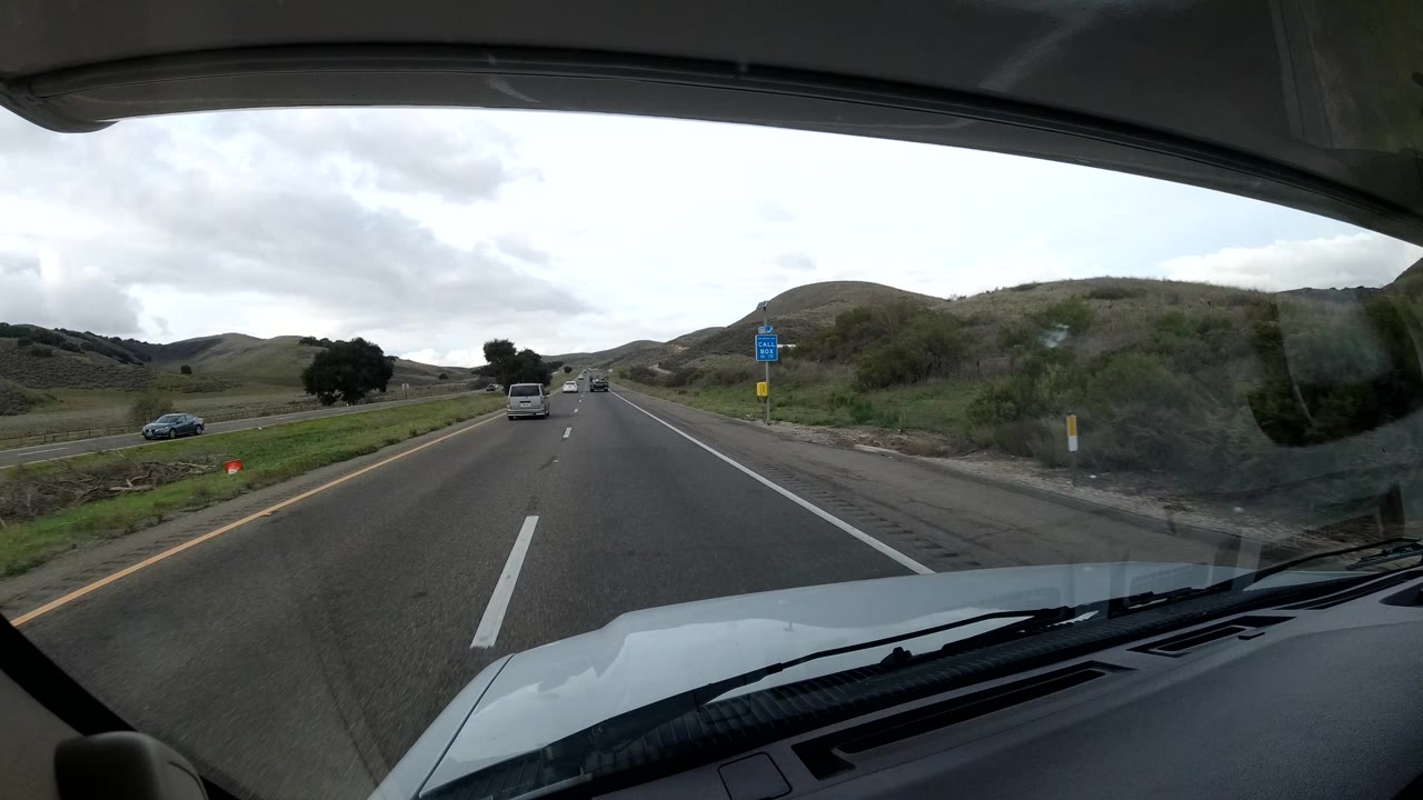 Forward dash view on a Central Coast California highway, with vehicles ahead, roadside signs to the right, rolling hills, cloudy light, and no visible people.