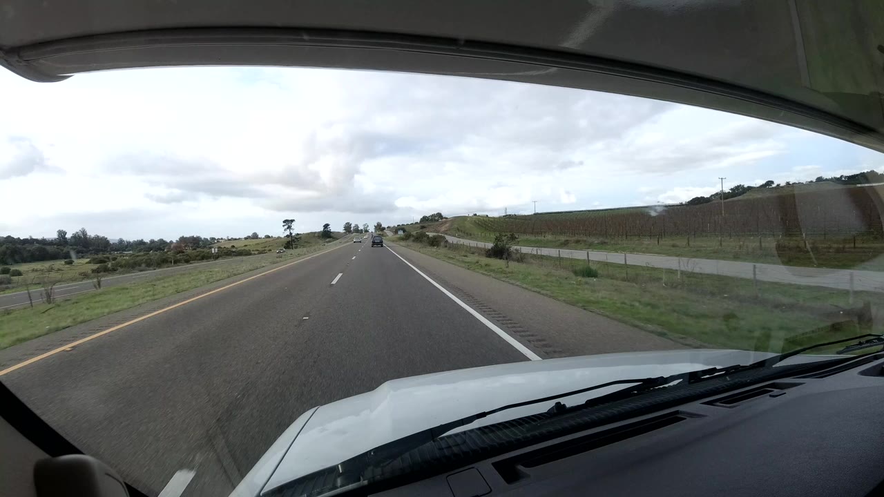 Forward vehicle POV on a California coast highway, with the white hood and windshield edge in the foreground, open lanes ahead, fields at the sides, cloudy sky, and no visible people.