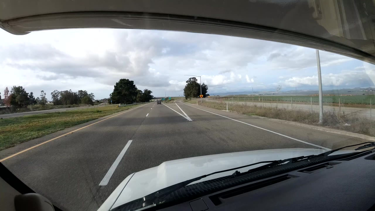 California coast vehicle POV near a ramp or lane split, with the hood low in frame, white road markings, open fields, roadside trees, cloudy sky, and no visible people.