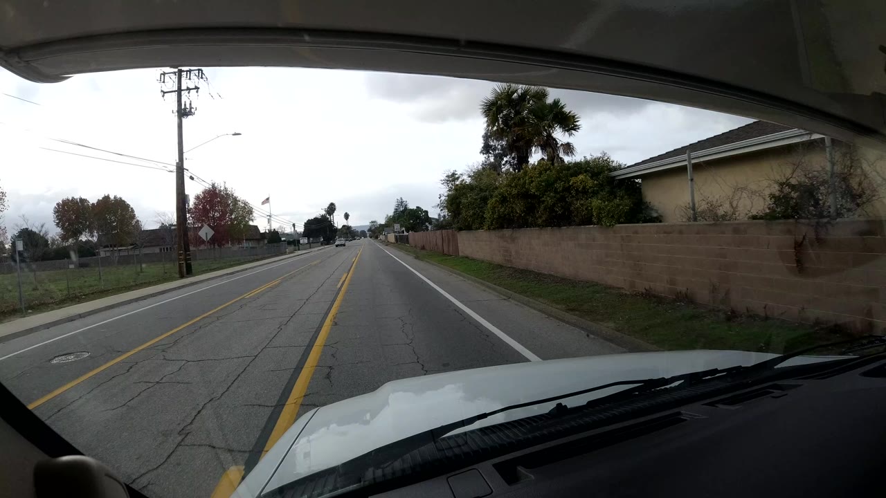 Forward-facing in-car view on a Coast, California residential road, with a white hood, windshield edge, sidewalks, block walls, homes, utility poles, trees, cloudy sky, and no visible people.