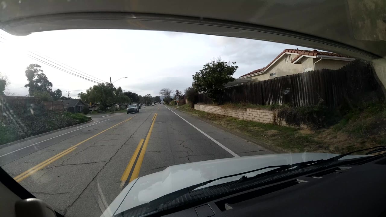 The in-car view in Coast, California passes homes, fences, sidewalk, shrubs, trees, overhead wires, and an oncoming vehicle on a two-lane road, with no visible people.