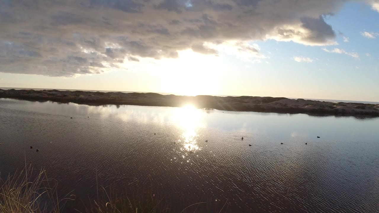 A bright low sun reflects on a lake in Pismo Beach, California, with dark shoreline hills, scattered clouds, foreground grasses, and no visible people.